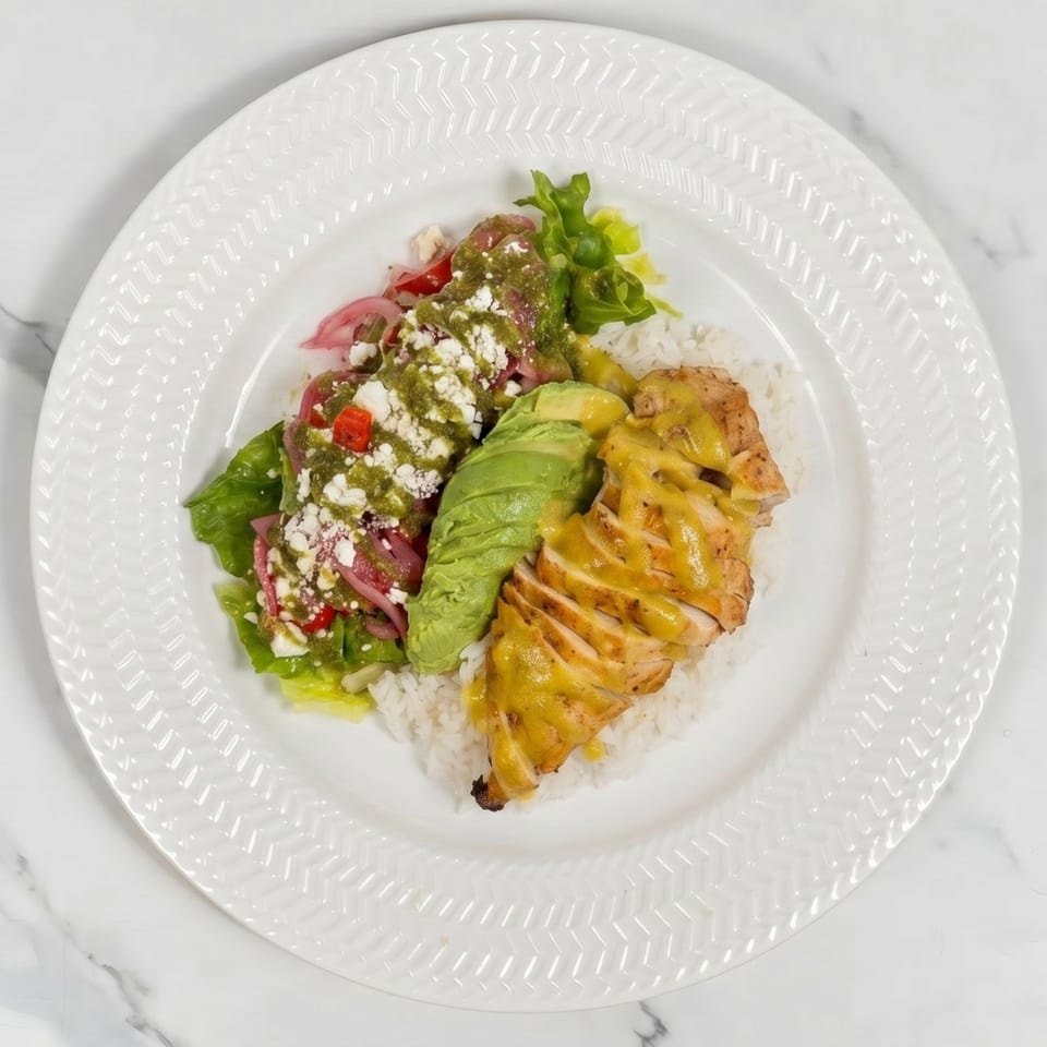 A white plate holds a chicken and rice bowl with avocado, salad, and green sauce.