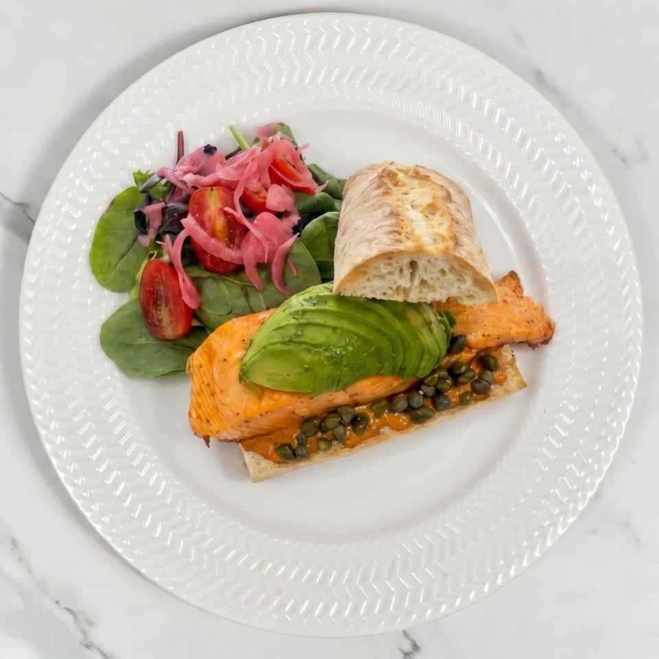 Salmon sandwich with avocado, capers, and a side salad with pickled onions and tomatoes.